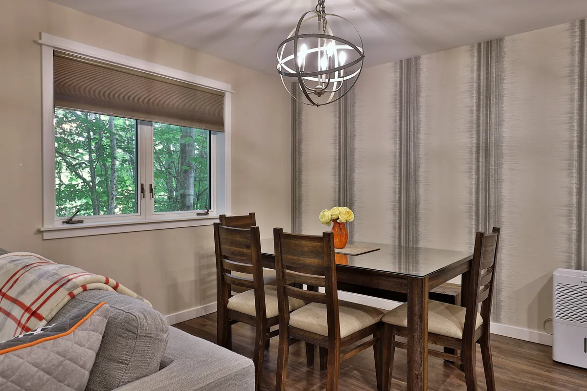 A warmly lit dining area featuring a wooden table adorned with a vase of bright yellow flowers. The space is enhanced by a striking chandelier above and a large window that looks out onto lush greenery, creating a seamless indoor-outdoor feel.