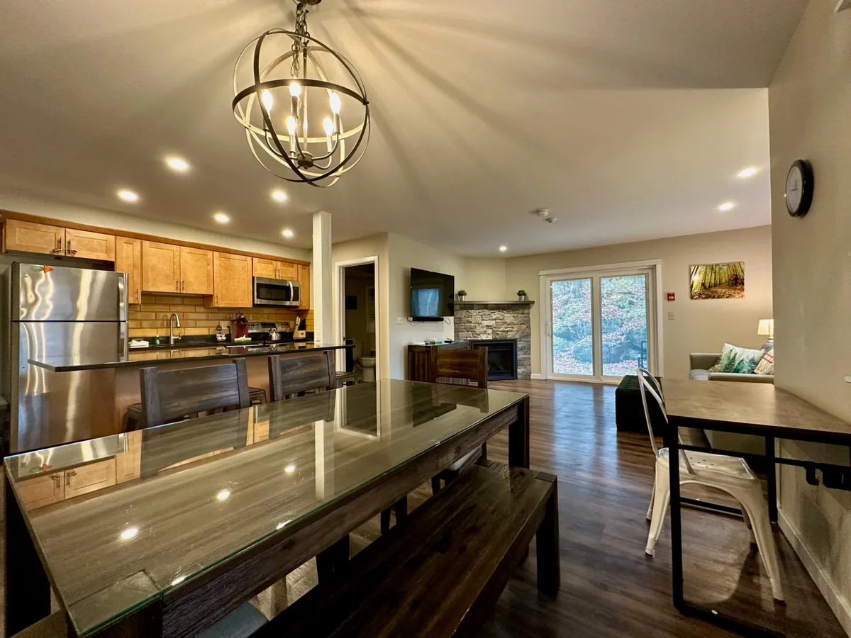 A welcoming dining area with a glass-top table, adjacent kitchen space, a fireplace, and large windows offering views outdoors. The overall atmosphere is warm, ideal for memorable group meals and relaxing evenings by the fire.