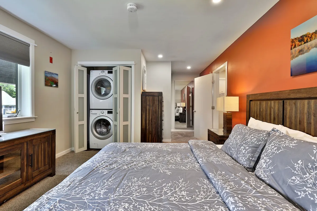 A cozy bedroom scene featuring a comfortable bed with floral bedding and a stacked washer and dryer tucked into the corner. Warm wooden furniture completes the inviting atmosphere, highlighting thoughtful in-unit conveniences.