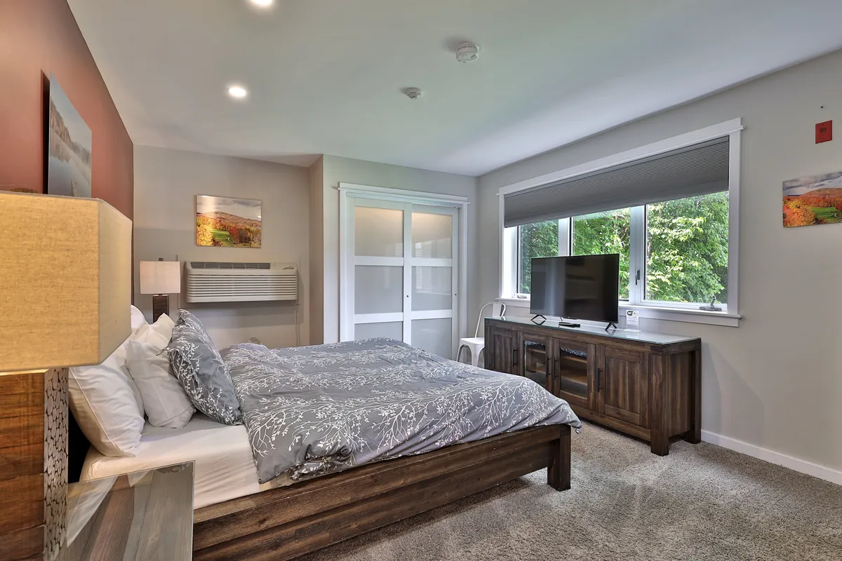 A bright, inviting bedroom featuring a bed with decorative bedding, a large window that brings in natural light, and classic wooden furniture. The arrangement is comfortable and spacious, offering a view out to the surrounding area, suggesting both privacy and openness for gatherings.