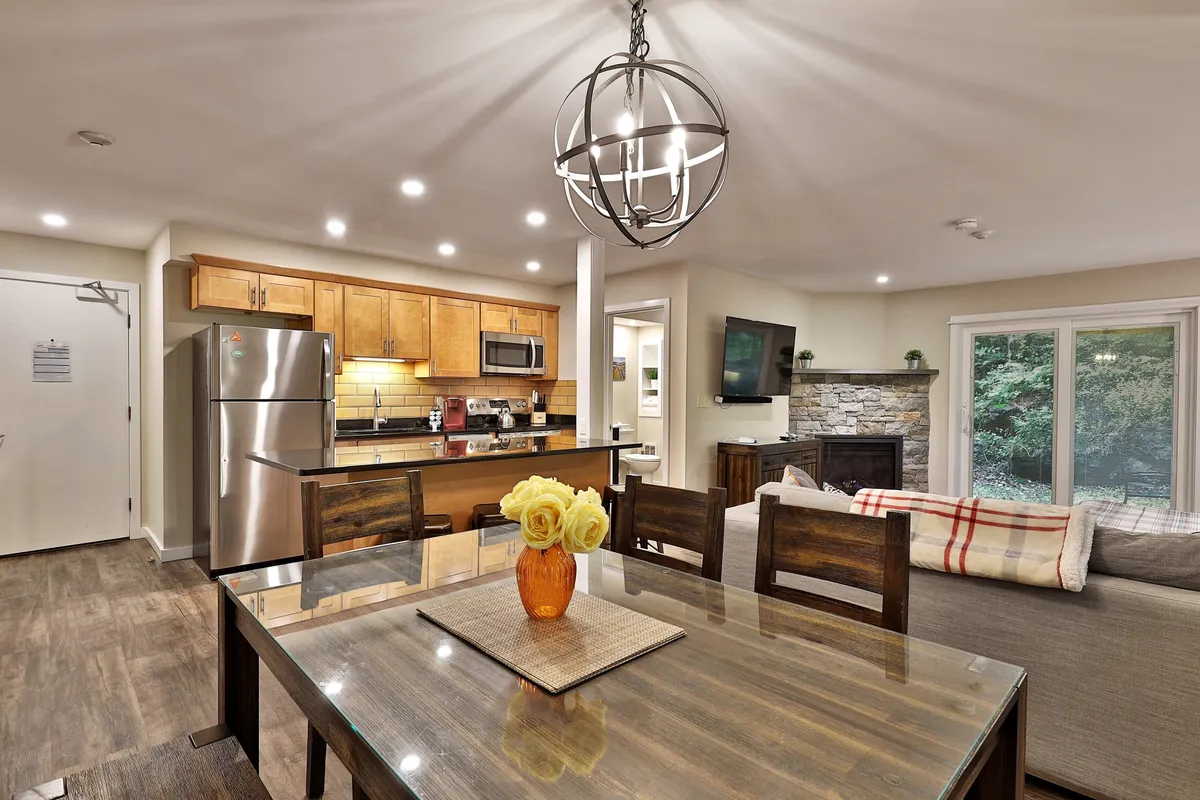 The image captures a stylish, open-plan living space featuring a modern dining table set for a meal, a sleek kitchen counter with stainless steel appliances, a warm fireplace, and a striking hanging light fixture overhead. The scene evokes a sense of comfort and togetherness, with inviting areas for cooking, dining, and relaxing by the fire.